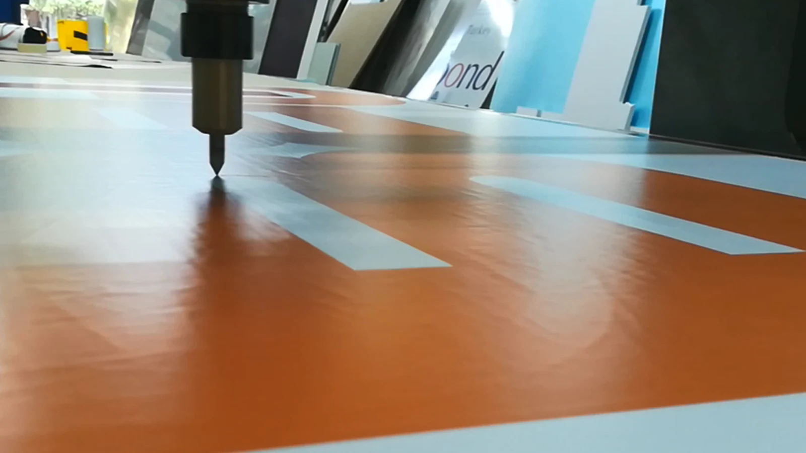A CNC blade cutting a contour in a printed sheet, panels stacked in the workshop behind